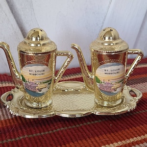 May include: A set of two gold-tone salt and pepper shakers with a vintage design. Each shaker features a colorful image of a steamboat with the text "St. Louis Missouri". The shakers are on a matching gold-tone tray.
