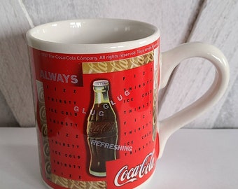 May include: White ceramic mug with a red Coca-Cola design. The design features a word search puzzle with the words "always", "fiz", "thirsty", "ice cold", "glug", and "refreshing". The mug also features a bottle of Coca-Cola.