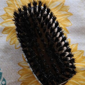 Peut inclure: Une brosse ovale noire et brune avec des poils noirs. La brosse est sur un fond floral blanc et jaune.