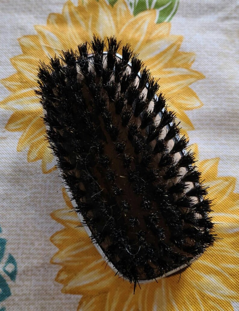 Peut inclure: Une brosse ovale noire et brune avec des poils noirs. La brosse est sur un fond floral blanc et jaune.