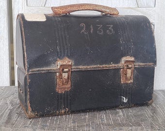May include: Vintage black metal lunchbox with a rusty handle and clasps. The number "2133" is handwritten on the front. The lunchbox shows signs of wear and tear, suggesting it is an antique item.