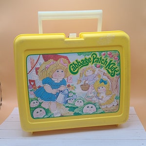May include: A yellow Cabbage Patch Kids lunchbox with a white handle. The front features a colorful illustration of Cabbage Patch Kids characters in a garden setting. The lunchbox is rectangular and has a latch closure.