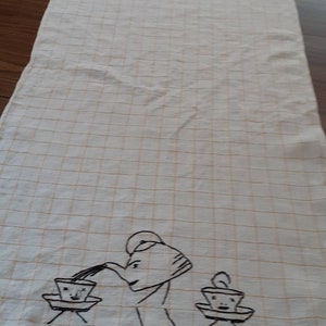 May include: A white tea towel with a grid pattern and a black embroidered design of a teapot pouring tea into two cups with faces.