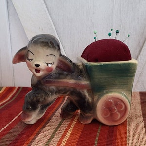 May include: A ceramic lamb figurine pulling a green and brown wagon with a red pincushion inside. The lamb has closed eyes and a pink nose.