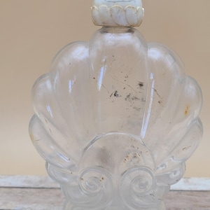 May include: A clear glass bottle with a carved shell design and a white, carved, round stopper. The bottle is made of clear quartz and has a slightly cloudy appearance.