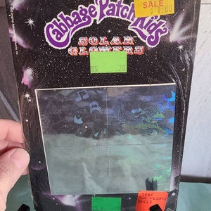 May include: A package of 3-D holographic stickers featuring the Cabbage Patch Kids Solar Glowers. The stickers are on a black background with stars and a comet. The package has a price tag that says "SALE $1.00".