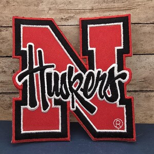 May include: A large embroidered patch in the shape of a block letter N, featuring a red center with a thick black and white border. The word Huskers is stitched in black script across the center, and a small registered trademark symbol is visible.
