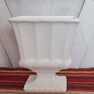 May include: White ceramic planter with a square top, a fluted body, and a pedestal base. The planter has a beaded trim around the top and base.