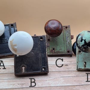 May include: Four antique door knobs and plates, each with a different style and color. The knobs are white, brown, black, and green. The plates are black, green, and brown.