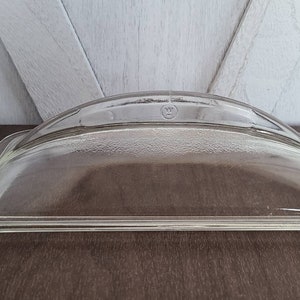 May include: Clear glass lid with a rounded top and a rectangular base. The lid has a raised edge around the perimeter and a small, raised, circular design in the center.