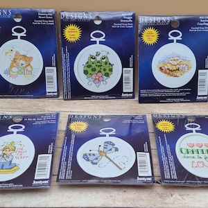 May include: Six counted cross-stitch kits. The kits feature designs of a cat, frog, seashell, girl praying, dragonfly, and text that reads "Grandma's home is full of love". Each kit includes colors and is packaged in a blue and white design.