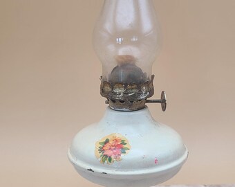 May include: A vintage white oil lamp with a clear glass chimney. The lamp features a floral decal and a metal wick adjuster. The base is round and the lamp is sitting on a wooden surface.