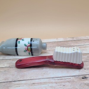 May include: A ceramic salt and pepper shaker set. One shaker is shaped like a tube with floral accents and black stripes. The other is a red handled brush with a white bristle top.