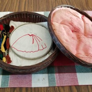 May include: A wicker basket with a lid, containing a white fabric with an embroidery hoop. The hoop has a red embroidered design of a skirt. The basket also contains several spools of embroidery floss in various colors.