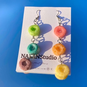 May include: Colorful dangle earrings featuring cereal-shaped charms in various colors, including green, pink, and yellow. The earrings are suspended from silver-tone hooks and connected by small chains. The earrings are displayed on a white card with the text "NANNSstudio".
