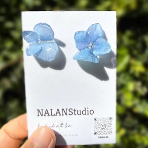 May include: A pair of light blue flower earrings. The earrings are made of a translucent material, with a small silver accent in the center of each flower. The earrings are displayed on a white card with the text "NALANStudio" and "handmade with love".