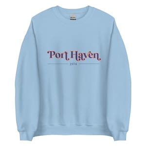 Könnte beinhalten: Hellblauer Pullover mit einem dunkelblauen und orangenfarbenen Aufdruck mit der Aufschrift "Port Haven 1974".