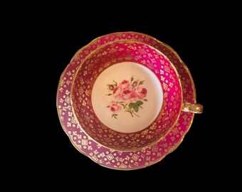 May include: A pink and gold teacup and saucer set with a floral design. The saucer has a scalloped edge.