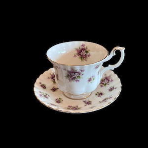 May include: A white teacup and saucer set with a floral pattern of purple and green violets. The set has a gold trim around the edges.