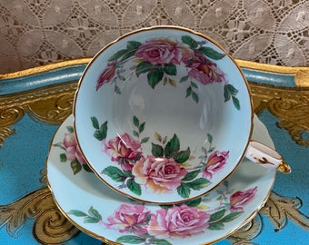 May include: A vintage teacup and saucer set with a delicate floral pattern featuring pink roses and green leaves on a light blue background. The set has a gold trim around the edges.