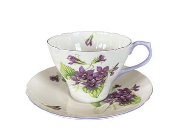 Könnte beinhalten: Ein weißes Tee-Set mit einer Tasse und Untertasse, verziert mit einem violetten Blumenmuster. Die Tasse hat einen hellvioletten Henkel und Rand, und die Untertasse hat einen passenden violetten Rand. Die Veilchen sind lila mit grünen Blättern.