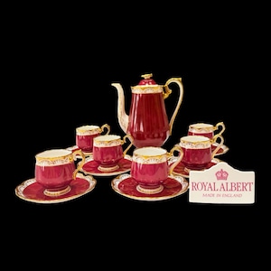 May include: A Royal Albert tea set featuring a deep burgundy teapot and six matching teacups with saucers. Each piece is accented with gold trim and white floral patterns. A white rectangular sign displays the text ROYAL ALBERT MADE IN ENGLAND.