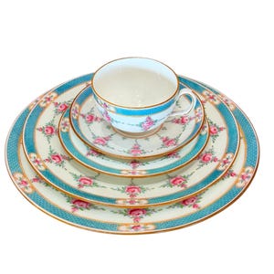 May include: A vintage tea set comprising a teacup and four plates. The set features a cream base adorned with pink rose floral patterns and teal blue borders. Gold trim accents the edges of the cup and plates, creating an elegant aesthetic.
