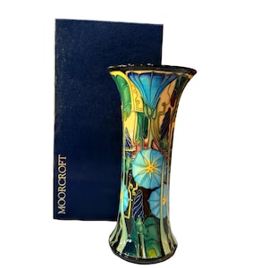 May include: A Moorcroft vase featuring a vibrant floral pattern with blue, yellow, and green tones on a cream background, accompanied by a dark blue rectangular box with the word MOORCROFT printed in white lettering on the side of the box.