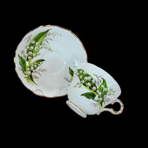 Puede incluir: Juego de taza y platillo blancos con un diseño floral de lirios de los valles. La taza y el platillo tienen un borde dorado. La taza tiene un asa curvada. El diseño floral presenta hojas verdes y flores blancas.