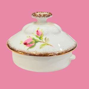 May include: A white ceramic trinket box with a lid, featuring a gold rim and a gold handle. The lid is decorated with pink rose buds and green leaves. The box has a round shape and is set against a pink background.