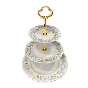 May include: A three-tiered white ceramic cake stand with a gold metal handle. The stand is decorated with a blue and green floral pattern.