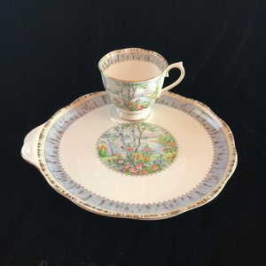Pu&ograve; includere: Un set vintage di tazza e piattino da t&egrave;. La tazza e il piattino sono bianchi con una scena paesaggistica con betulle, fiori e un lago. Il piattino ha un bordo azzurro con finiture dorate. La tazza ha un manico e finiture dorate.