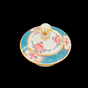 May include: A round, white ceramic lid with a floral design. The lid features a teal border with a scale pattern, a gold trim, and pink roses. A white knob with gold accents sits atop the lid.