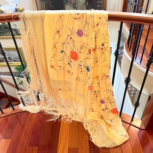 May include: A pale yellow rectangular shawl featuring intricate floral embroidery in shades of orange, purple, and blue. The fabric is adorned with delicate vine patterns and finished with long, knotted fringe along the edges, creating a textured look.