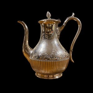 Może przedstawiać: Srebrny czajnik z ozdobnym wzorem i karbowanym dnem. Czajnik ma pokrywkę z gałką i zakrzywiony uchwyt.