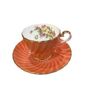 May include: A coral coloured teacup and saucer set with a floral design. The cup and saucer have a gold trim.