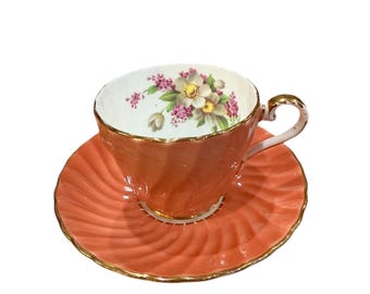 May include: A coral colored teacup and saucer set with a floral design. The cup and saucer have a gold trim.