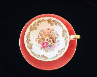 May include: A vintage teacup and saucer set with a floral pattern. The cup and saucer are both white with a gold rim and a floral design in shades of pink, blue, and green. The saucer is a bright coral color.