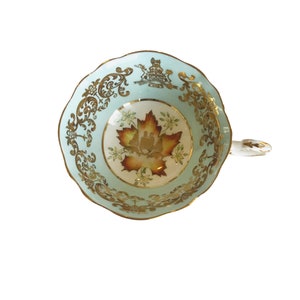 May include: A light blue teacup with a gold rim and intricate gold floral designs. The centre of the cup features a painted orange and yellow leaf with a gold crest.