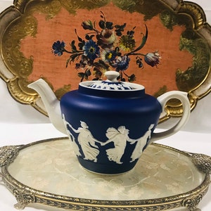 Puede incluir: Una tetera azul y blanca con un diseño blanco de figuras bailando en el lateral. La tetera está sentada en una bandeja dorada y blanca con un diseño floral.