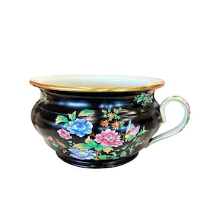 May include: A black ceramic planter with a gold rim and a handle. The planter is decorated with colourful floral and butterfly designs in pink, blue, and green. The interior is white.