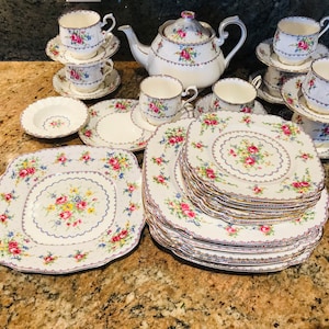 May include: A set of vintage floral teacups and saucers with a teapot and a variety of plates. The teacups and saucers have a delicate floral pattern with gold accents. The teapot is white with a floral design and a gold lid. The plates are white with a floral pattern and a gold rim.