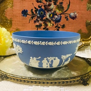 Może przedstawiać: Jasnoniebieska ceramiczna miska z białym wzorem kwiatowym wokół krawędzi i białą sceną z ludźmi w krajobrazie z przodu. Miska stoi na złotej tacy z kwiatowym wzorem.