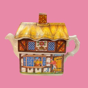 Puede incluir: Una tetera de cerámica con forma de cabaña, techo de paja amarillo y chimenea. El exterior presenta una escena pintada con una persona en una ventana y otra junto a una puerta, junto a un pequeño perro azul y detalles de motivos florales.