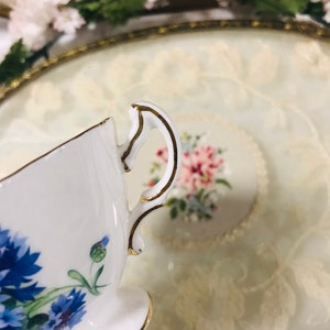 May include: A white teacup with a gold rim and a floral design featuring blue flowers. The teacup is sitting on a white saucer with a gold rim.