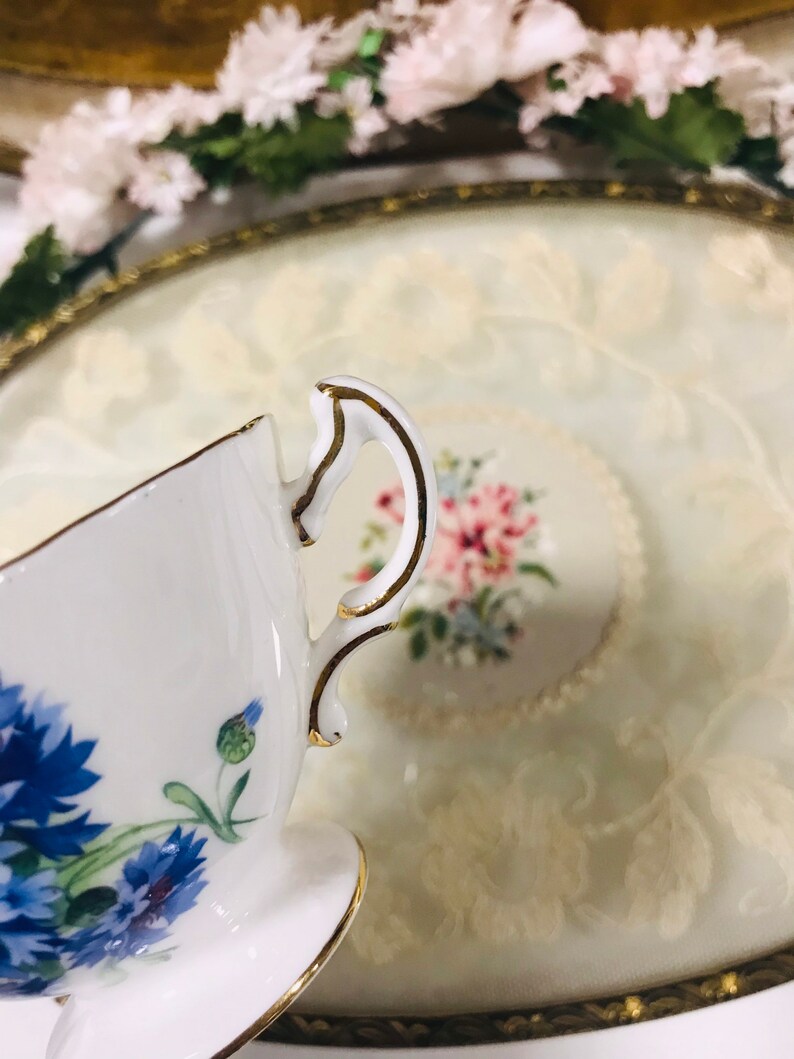 May include: A white teacup with a gold rim and a floral design featuring blue flowers. The teacup is sitting on a white saucer with a gold rim.