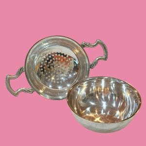 May include: A polished silver-toned metal strainer with two ornate, curved side handles and a perforated base, positioned alongside a matching polished silver-toned metal bowl with a smooth, rounded interior and a plain, circular rim.