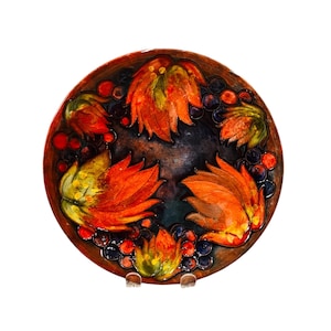 May include: A round ceramic plate with a colourful floral design. The plate features a repeating pattern of orange, yellow, and green flowers with blue berries. The plate has a dark brown background.