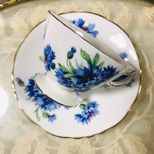 May include: A white teacup and saucer with a blue and green floral pattern. The cup and saucer have a gold trim.