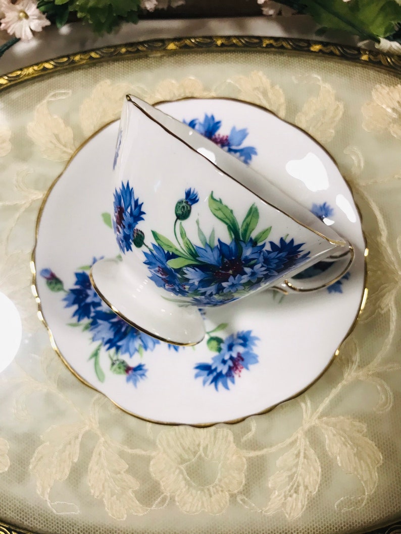 May include: A white teacup and saucer with a blue and green floral pattern. The cup and saucer have a gold trim.